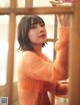 A woman in an orange sweater leaning against a window.